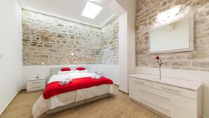 Une chambre avec un mur en pierre, un lit avec une literie rouge et blanche, une commode blanche et un miroir.