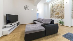 Un salon moderne avec un téléviseur à écran plat posé sur un meuble blanc, un canapé gris, un mur en pierre et une lucarne.
