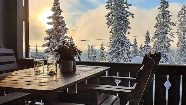 Un ponte di legno con tavolo e sedie, con vista su una foresta innevata e alberi sempreverdi alti.