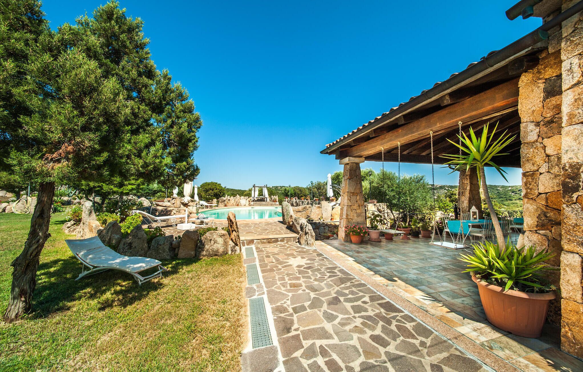 A stone pathway leading to a pool area with lounge chairs and a pergola with hanging plants.