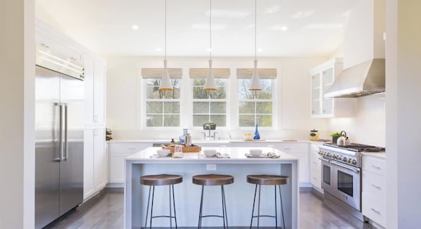 Una cocina moderna con una isla central, taburetes de bar, electrodomésticos de acero inoxidable y ventanas amplias.