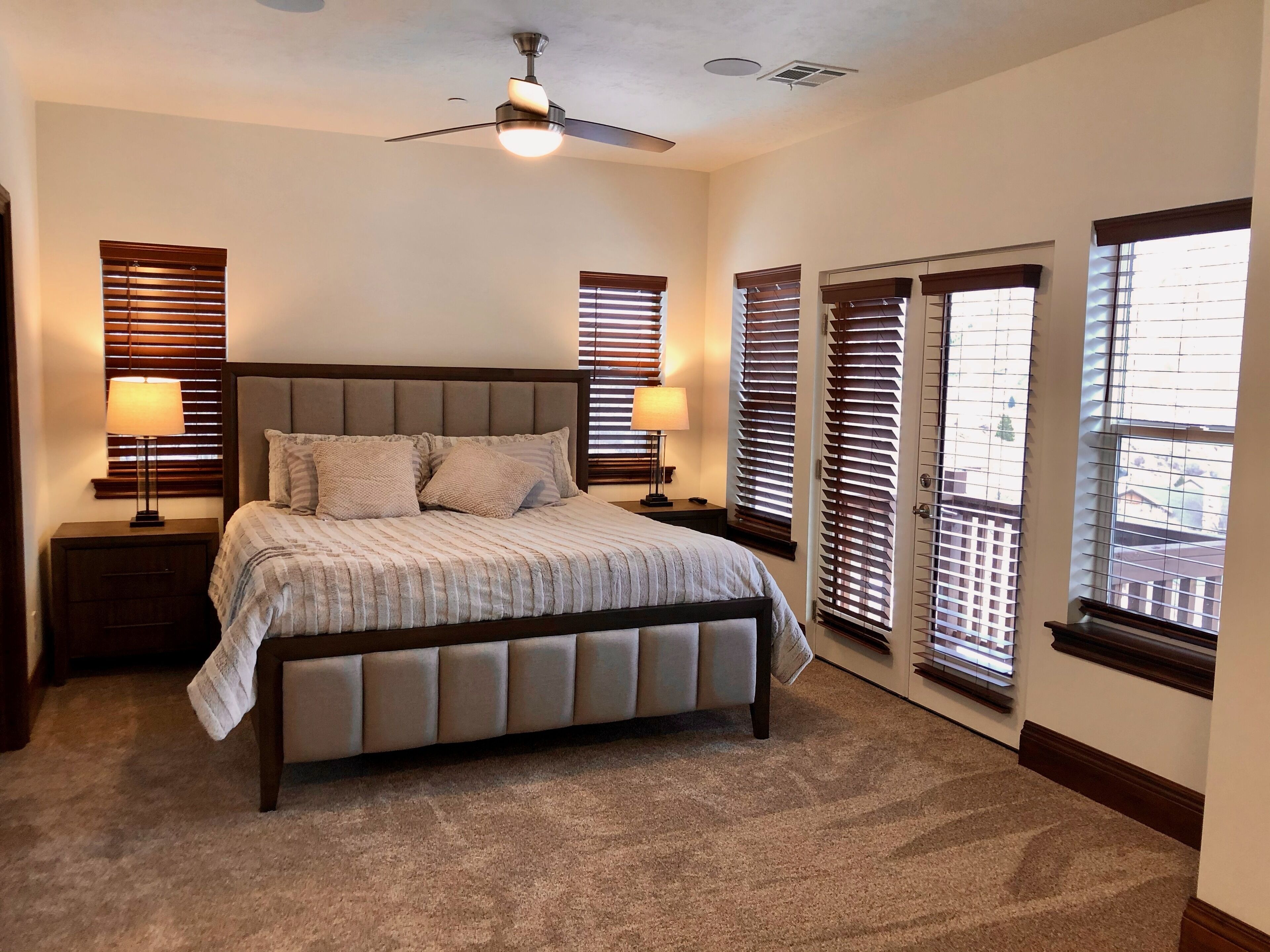 A bedroom with a bed, two bedside lamps, a ceiling fan, and multiple windows with blinds.