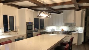 Uma cozinha moderna com armários brancos, uma ilha grande com banquetas e forno embutido, além de uma luminária pendente.