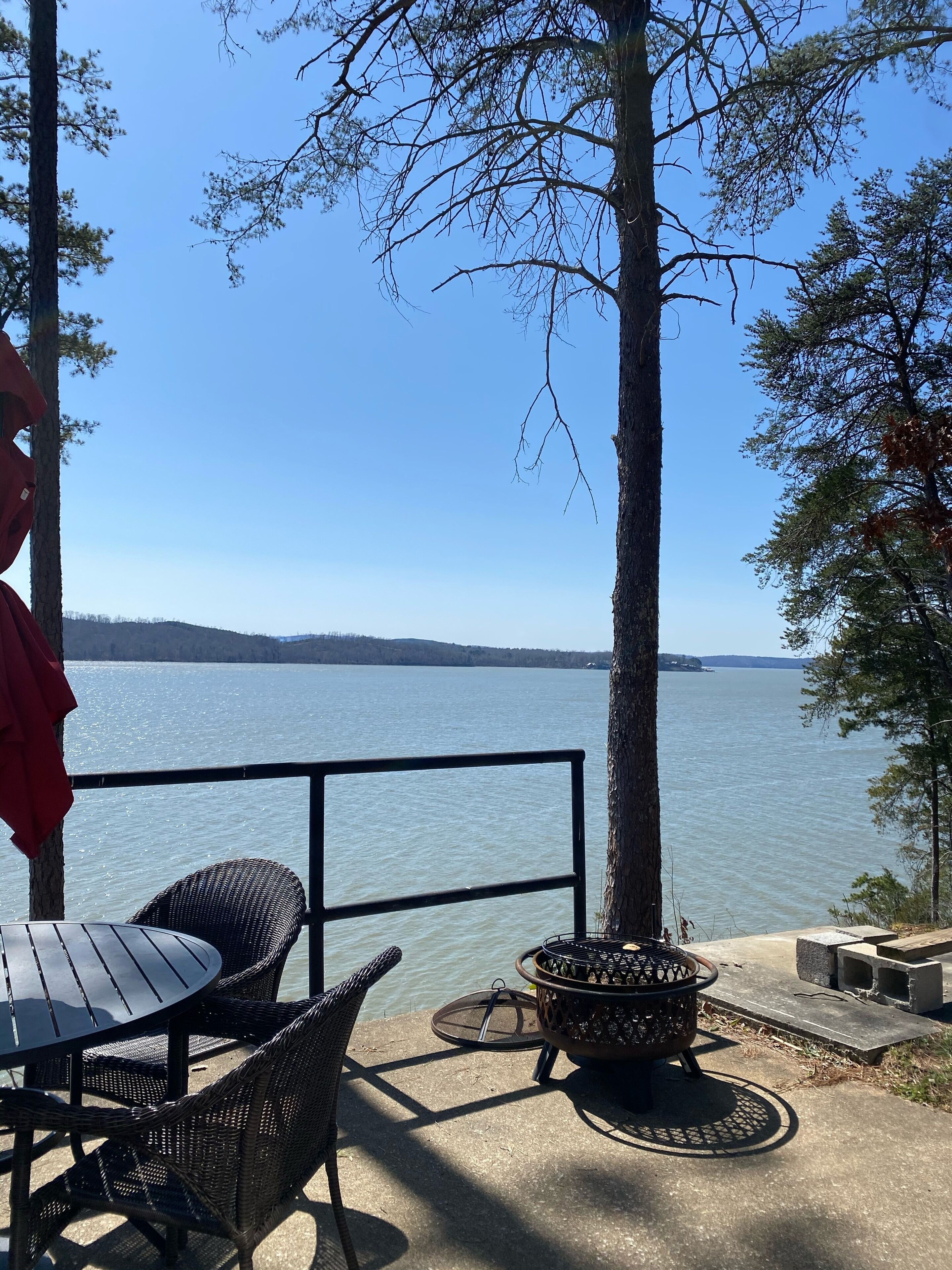 A patio with wicker chairs and a table overlooking a lake, with a fire pit and a red umbrella.