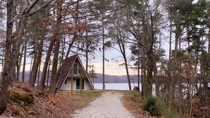 Eine Hütte mit einem Dreiecksdach, umgeben von hohen Bäumen und einem See im Hintergrund.