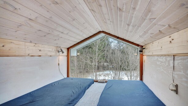 Una habitación con techo inclinado, una cama con colchón azul y una ventana con vista a árboles.