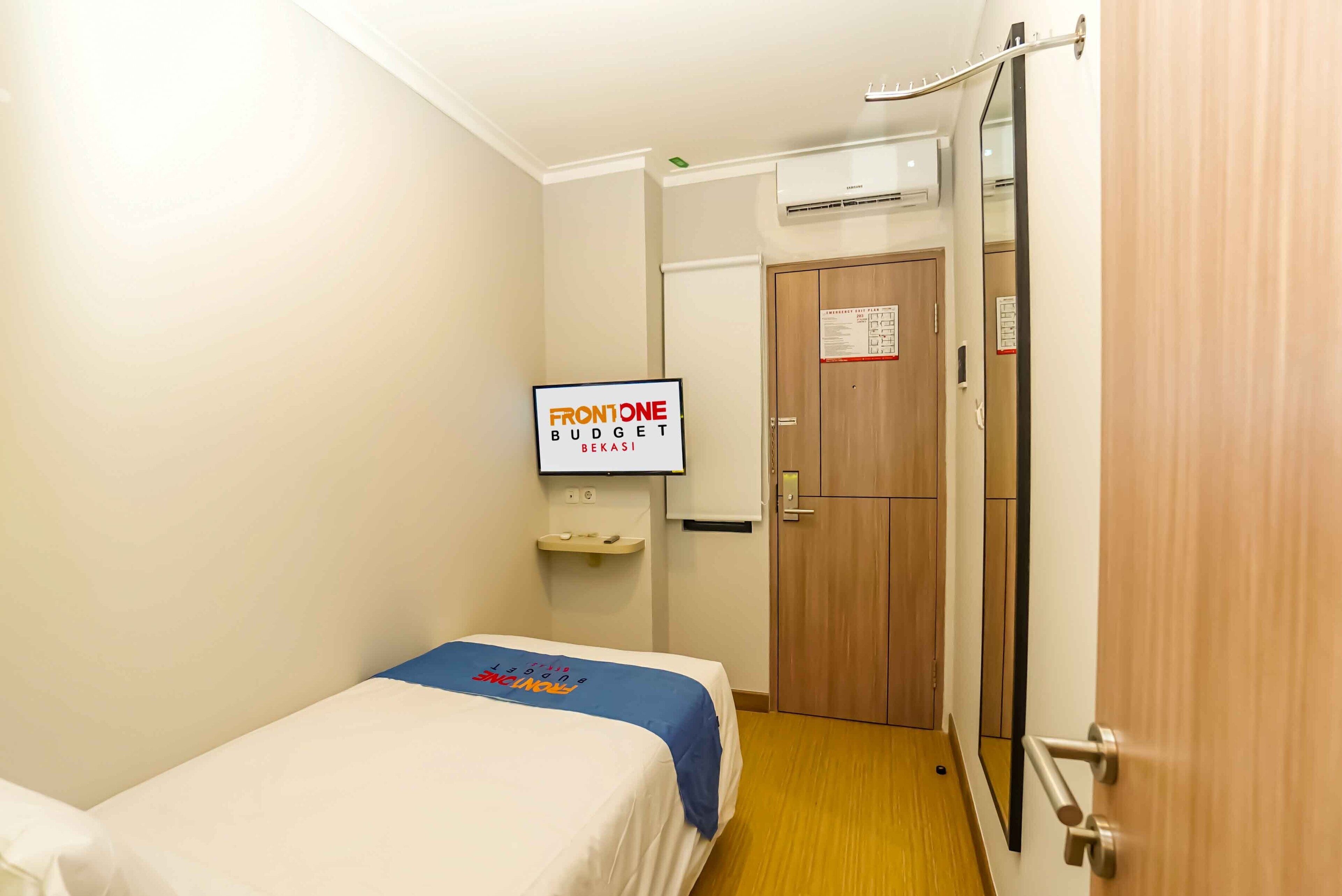 A hotel room with a bed, a television displaying 'FRONTONE BUDGET BEKASI', a wooden door, and an air conditioning unit.