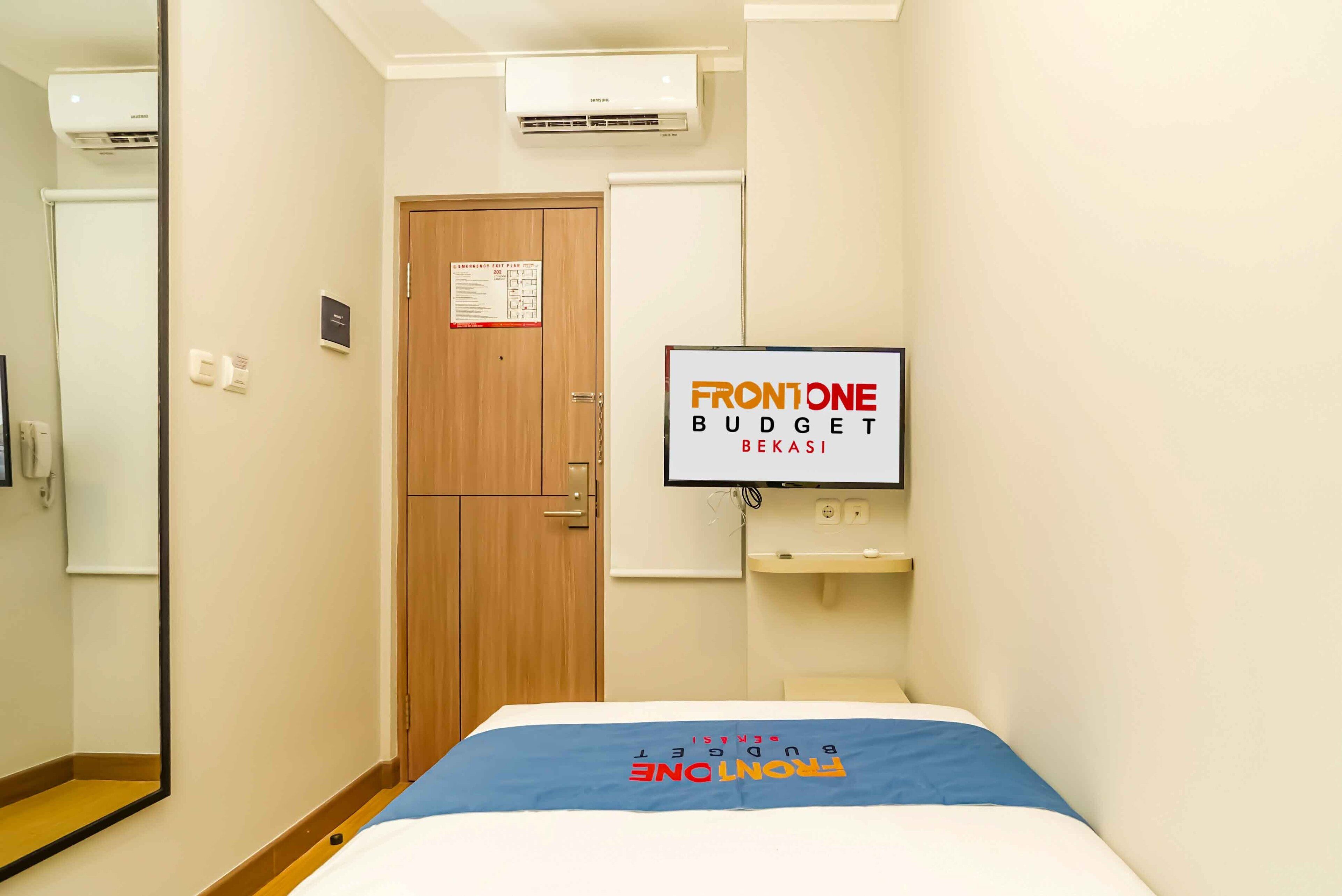 A hotel room with a bed, a wooden door, a wall-mounted TV, a small sink, and a mirror.