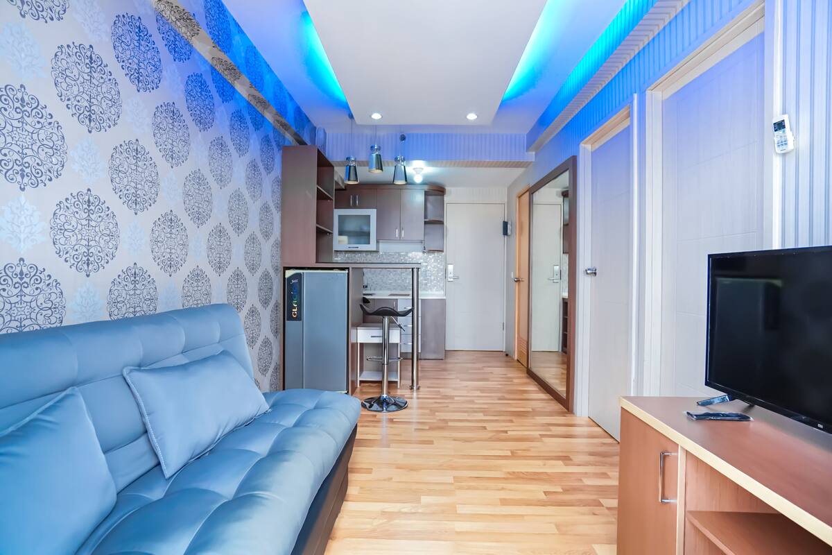 A modern living room with a blue sofa, wooden flooring, a TV, and a kitchen area in the background.