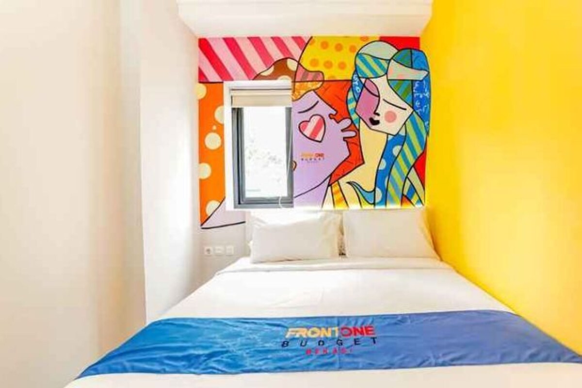 Una cama con un mural colorido en la pared, una ventana y una colcha azul con el texto 'FRONTONE BUDGET'.