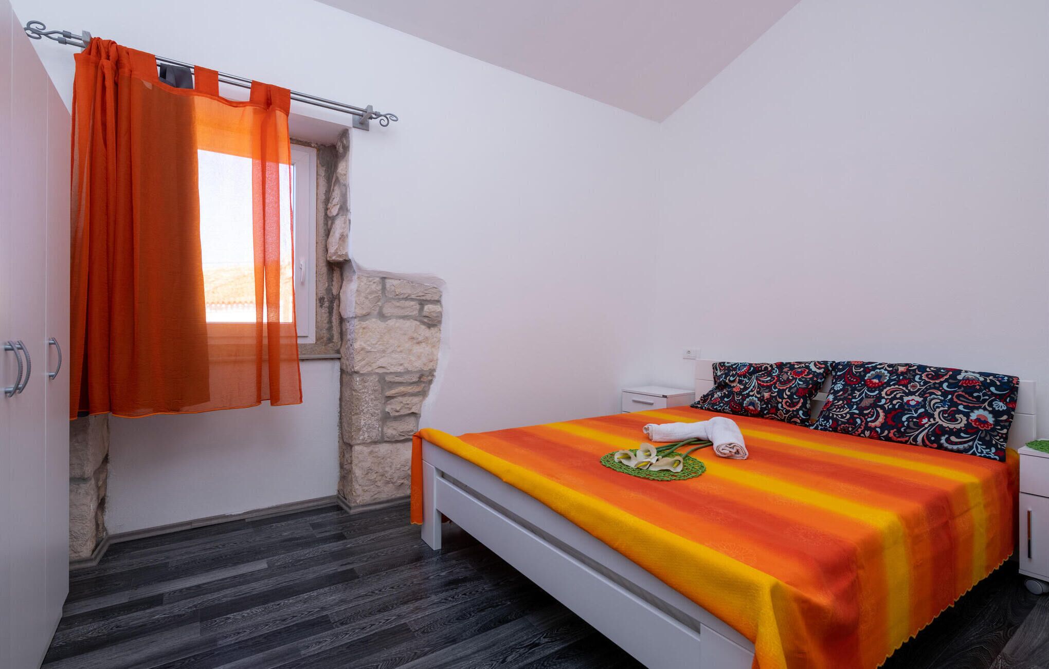 Un dormitorio con cama, mesita de noche, una pequeña mesa, ventana con cortinas naranjas y pared de piedra.