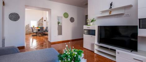 A modern living room with a flat-screen TV, a grey sofa, and a dining area with a table and chairs.