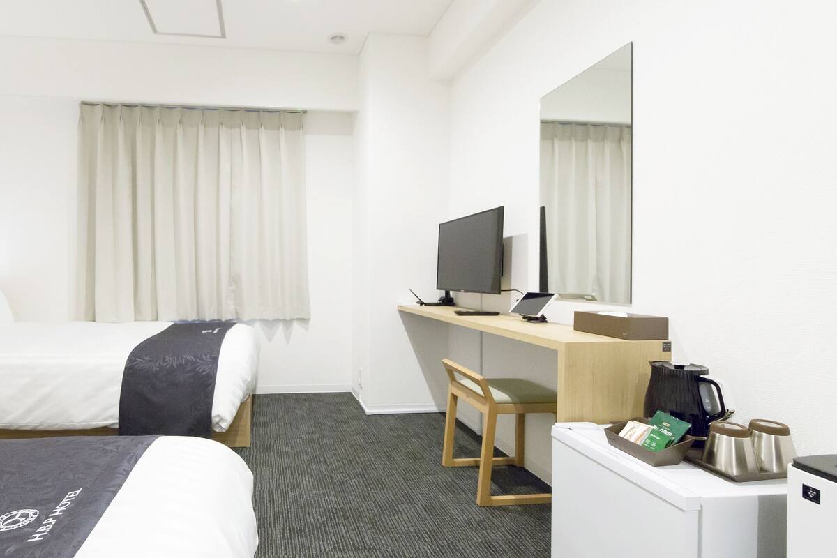 Habitación de hotel con dos camas, un escritorio con ordenador, una silla, cafetera y un refrigerador pequeño.
