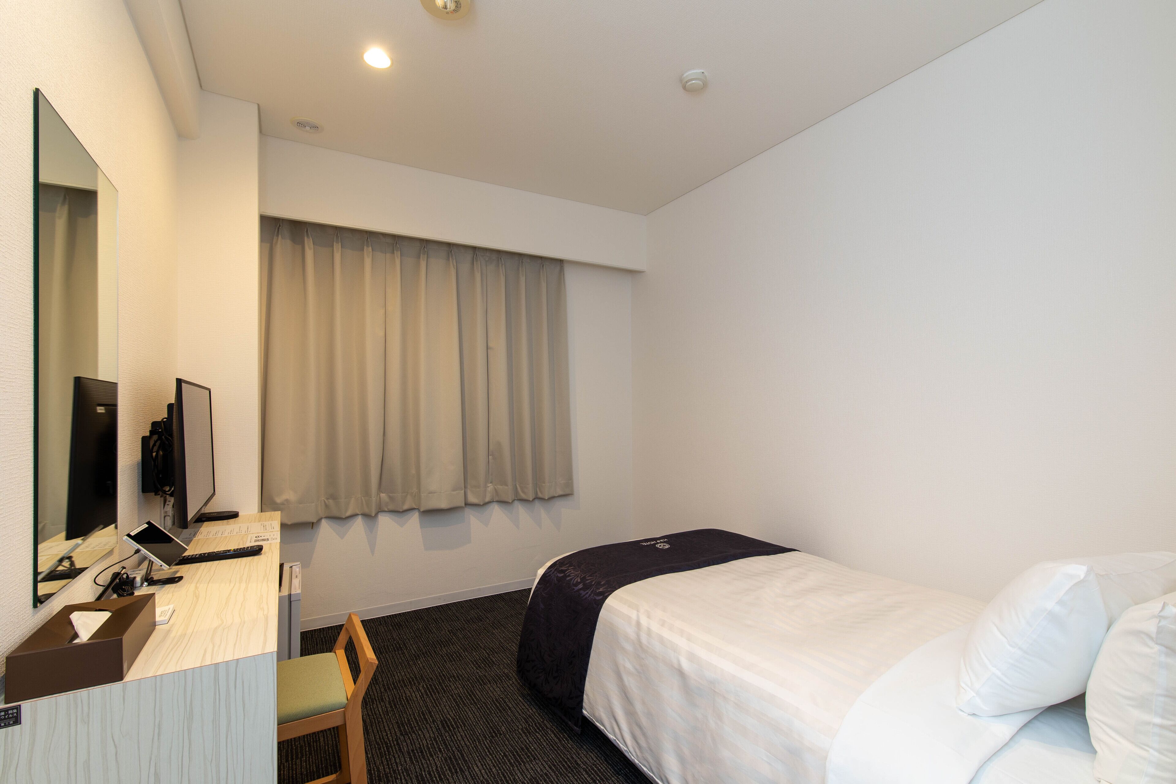 A hotel room with a bed, a desk with a computer, a chair, a large mirror, and a window with curtains.