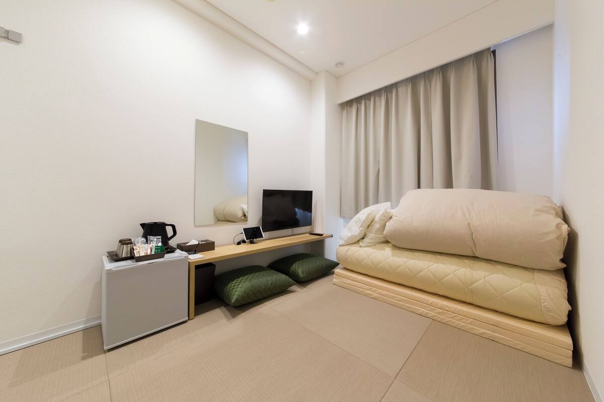 Una habitación de hotel moderna con cama, escritorio con televisor, refrigerador pequeño y ventana con cortinas.
