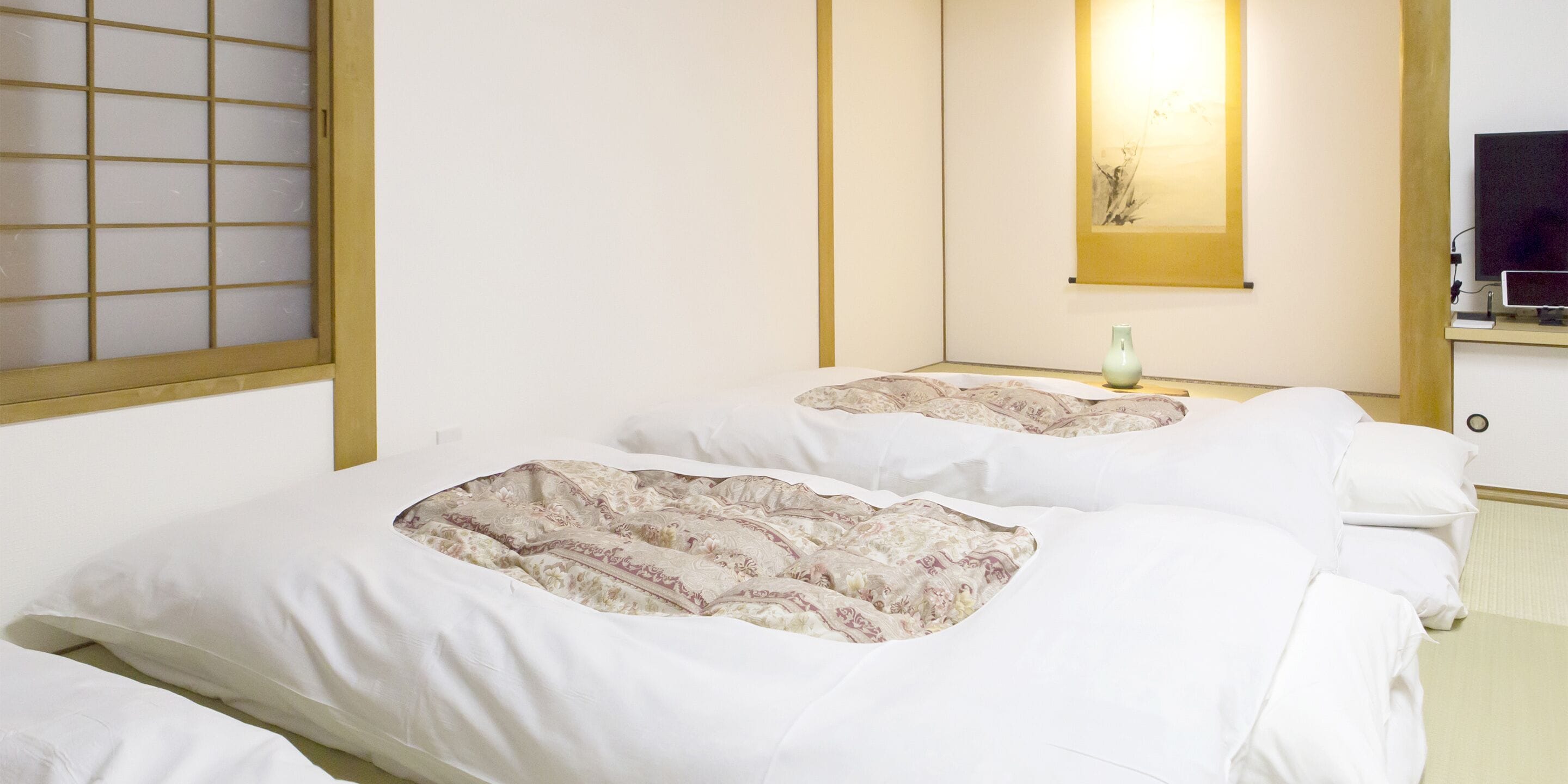 Habitación de hotel con dos camas, una mesita de noche con un florero, un televisor montado en la pared y un ventanal con diseño de cuadrícula.