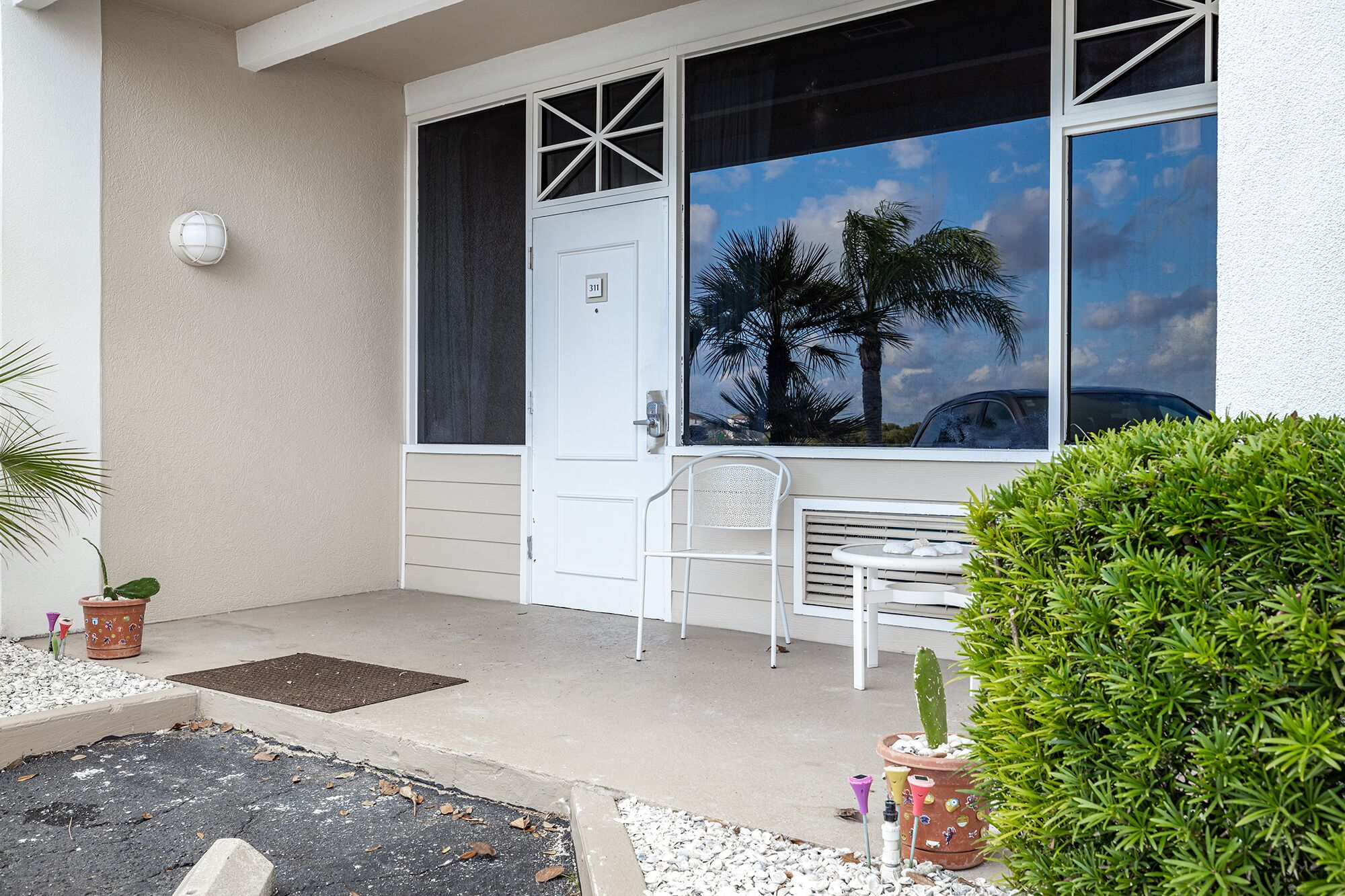 Vista frontal de una casa con una puerta blanca, una ventana que da a palmeras, una silla blanca y plantas en macetas.