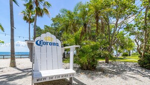 A wooden sign with "Corona" and "Where the fun never sets" text, surrounded by palm trees and a beach view.