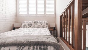 A bedroom with a bed, two bedside tables, a window with shutters, and a wooden railing. - BAY PROPERTIES | Pippi Beach Studio (Yamba)