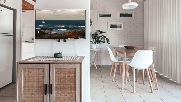 A modern kitchen with a large TV, white cabinets, and a dining area with a table and chairs. - BAY PROPERTIES | Pippi Beach Studio (Yamba)