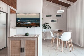 A modern kitchen with a large TV, white cabinets, and a dining area with a table and chairs. - BAY PROPERTIES | Pippi Beach Studio (Yamba)