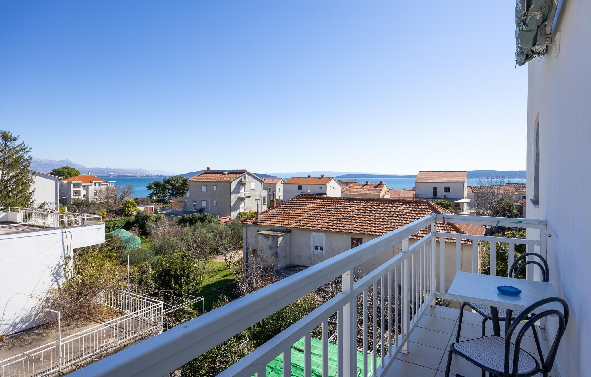 A balcony with a table and chairs, overlooking a coastal town with terracotta-roofed buildings and a clear blue sky.