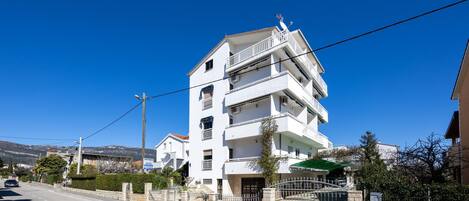 A multi-story white building with balconies, a gated entrance, and a clear blue sky.