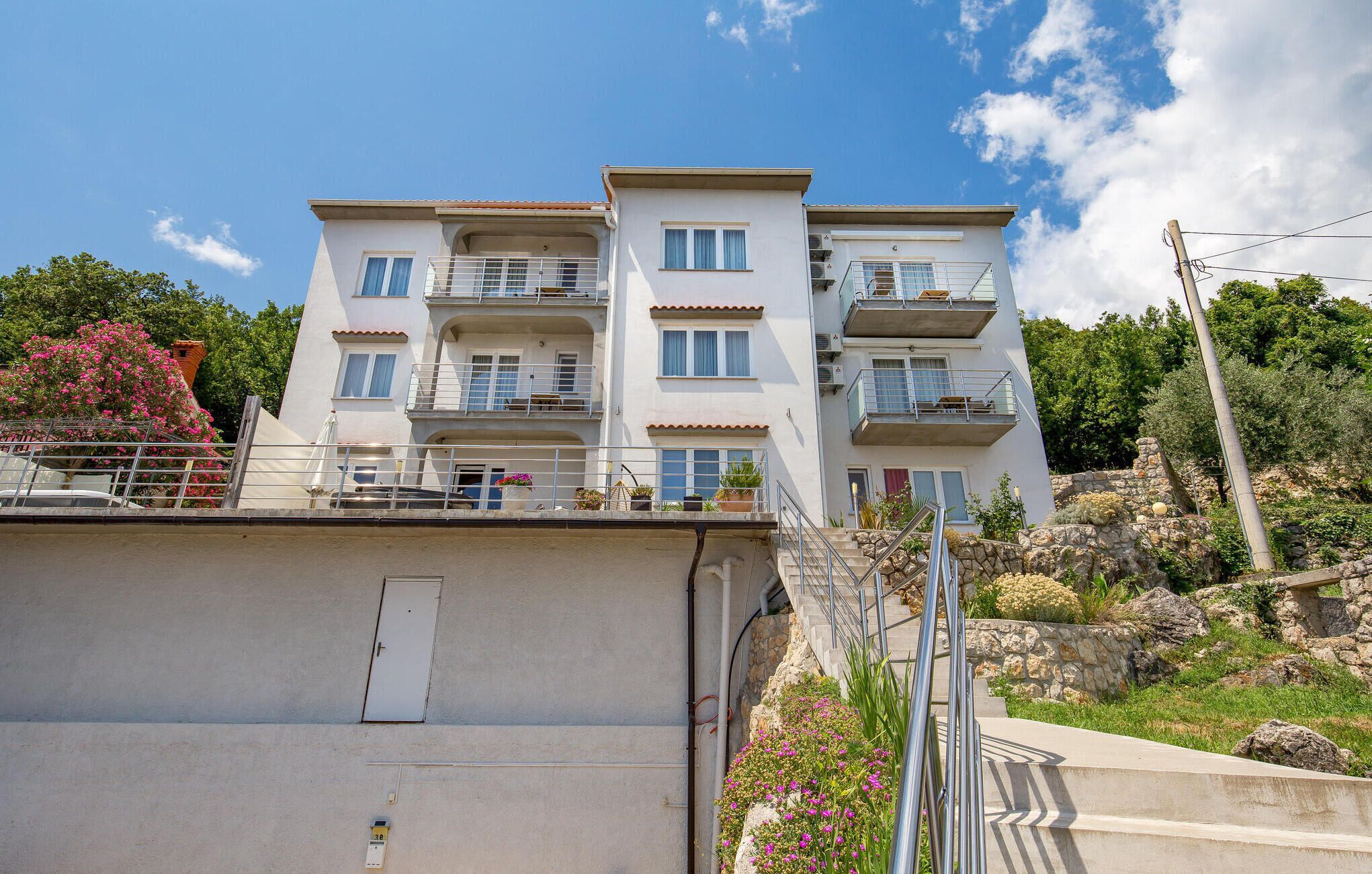 A multi-story residential building with balconies, a stone facade, and a garden area with pink flowers.