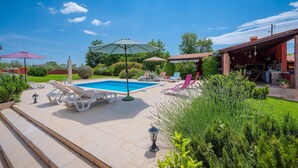 Un espace piscine agrémenté de chaises longues et de parasols, entouré de verdure et bordé d’un bâtiment au toit rouge.