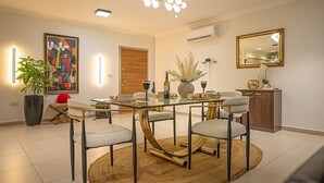 A dining area with a glass table, four chairs, a woven rug, a potted plant, a painting, and a mirror on the wall. - Spacious 2BR Penthouse Apt close to Airport with pool & gym (Accra)