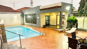 A modern outdoor pool area with a clear pool, a covered seating area, and a staircase leading to an upper level. - Spacious 2BR Penthouse Apt close to Airport with pool & gym (Accra)