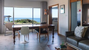 A modern living room with a dining area, large windows with a view of the ocean, a ceiling fan, and a wooden accent wall. - BAY PROPERTIES | Ocean Sounds (Yamba)