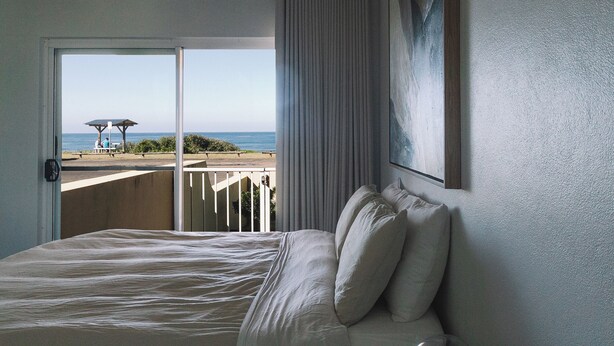 A bedroom with a large bed, a view of the beach, and a balcony with a table and umbrella. - BAY PROPERTIES | Ocean Sounds (Yamba)