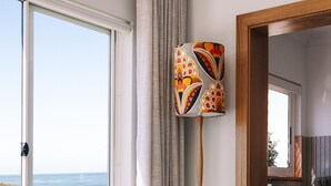 A modern living room with a large window, a grey cushioned chair, a colorful abstract painting, and a wooden crate. - BAY PROPERTIES | Ocean Sounds (Yamba)