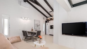 Una sala moderna con un sofá beige, una mesa de comedor con sillas, una pequeña mesa blanca con un jarrón de flores, un televisor de pantalla plana montado en la pared y una cocina con armarios blancos y un microondas.