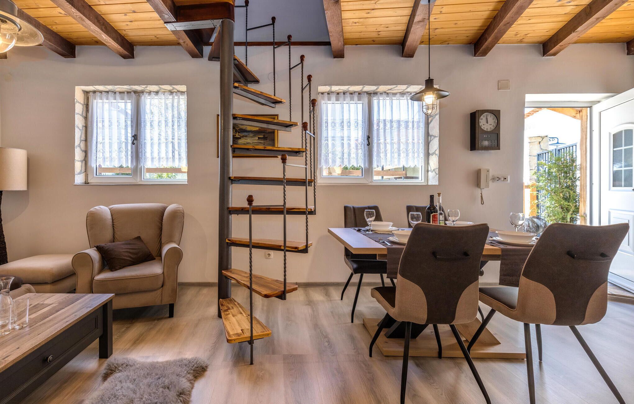 Una sala de estar acogedora con un sofá, una mesa de comedor con sillas, una escalera de madera y vistas al exterior a través de amplios ventanales.