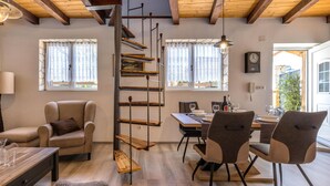 Un salon chaleureux avec un canapé, une table à manger et des chaises, un escalier en bois et une vue sur l’extérieur grâce à de grandes fenêtres.