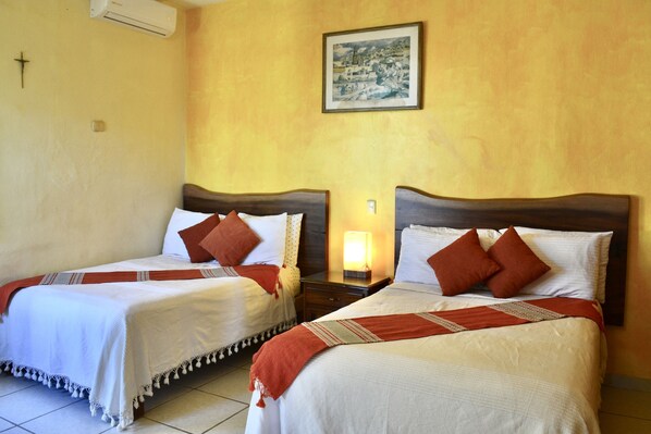Two single beds with wooden headboards, white bedding, and red throw pillows. There is a wall-mounted air conditioner and a framed picture above the beds.