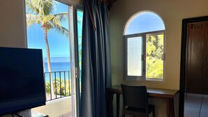A room with a TV on a wooden cabinet, a desk with a chair, a window with a view of palm trees and the sea, and a door with a wooden frame. - Beachfront Villa Casa Mar de Grecia 2 (Punta de mita)
