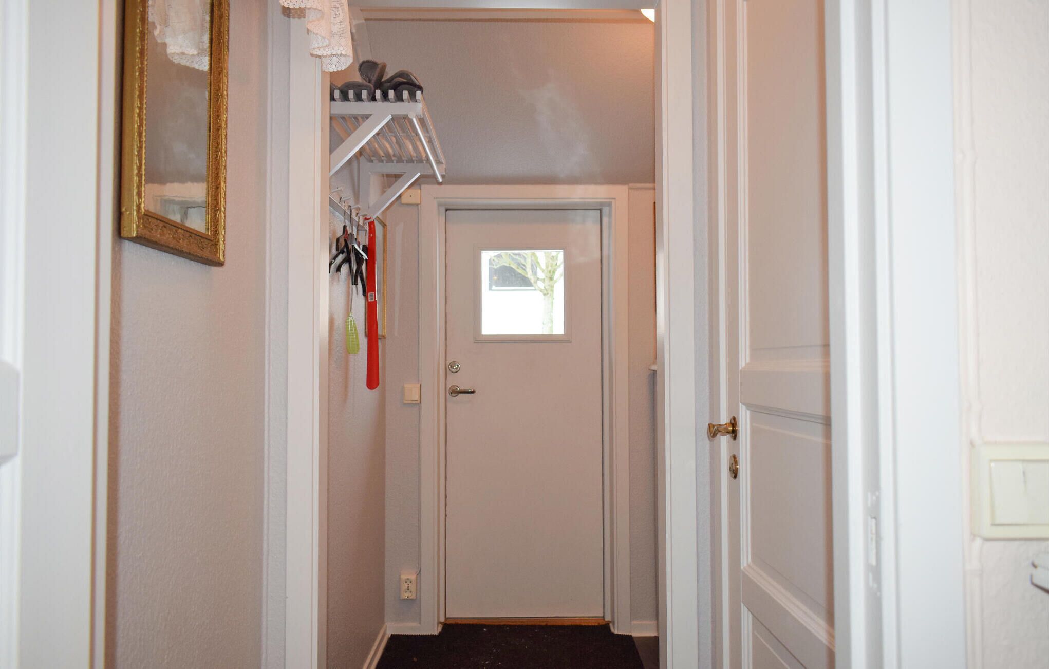 A narrow hallway with a white door at the end, a small window, and a coat rack with items above it.