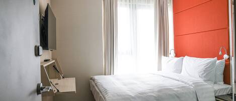 A hotel room with a bed, a desk, a chair, a TV, and a window with curtains.