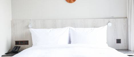 Una cama bien tendida con ropa de cama blanca, cabecera de madera y mesita de noche con un teléfono.