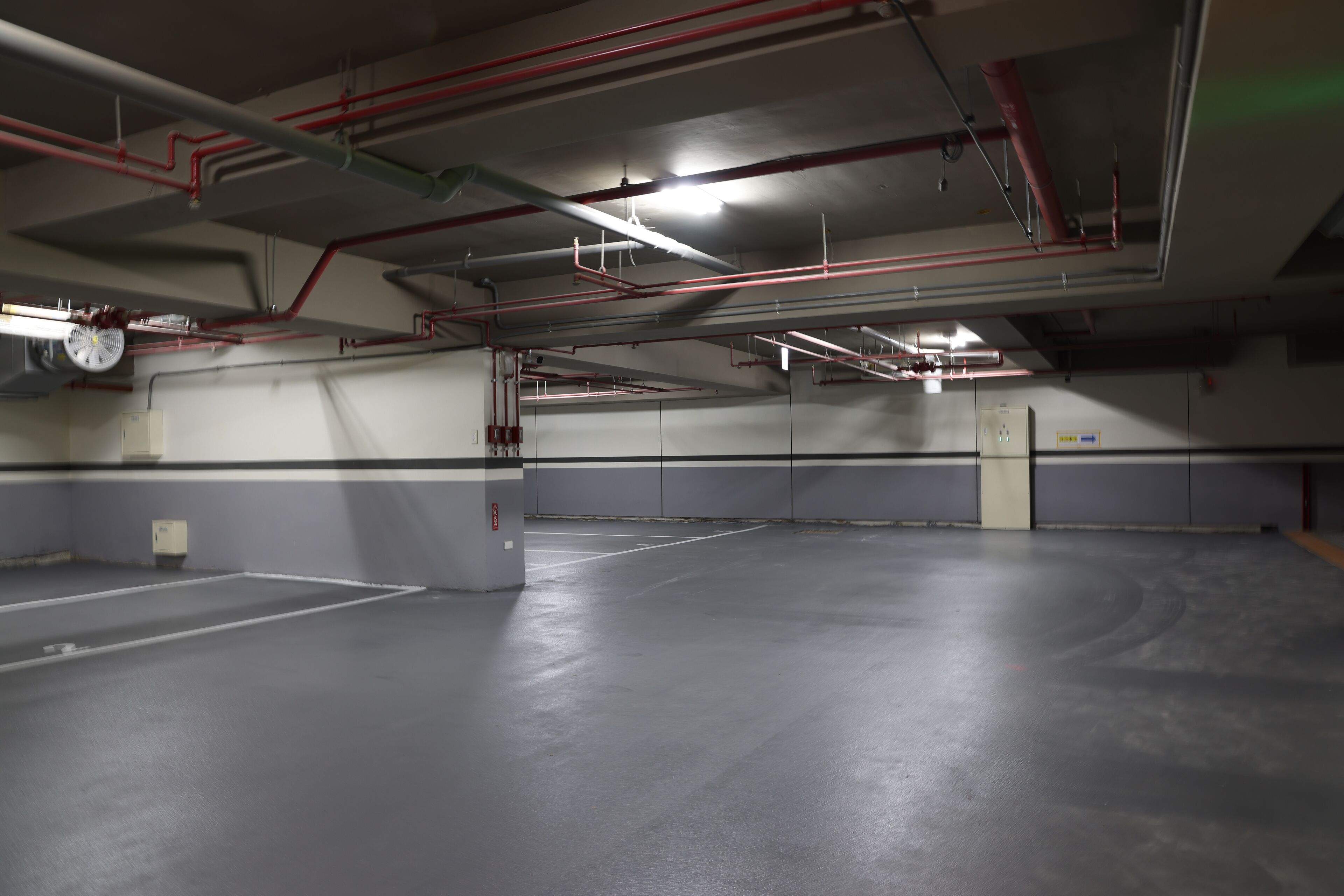 Een lege parkeergarage met grijze muren en betonnen vloeren, met zichtbare leidingen en een plafondventilator.