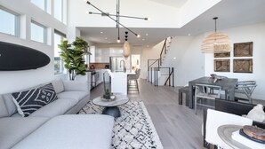 A modern living room with a sofa, a dining area with a table and chairs, a kitchen with white cabinets, and a staircase leading to another level.