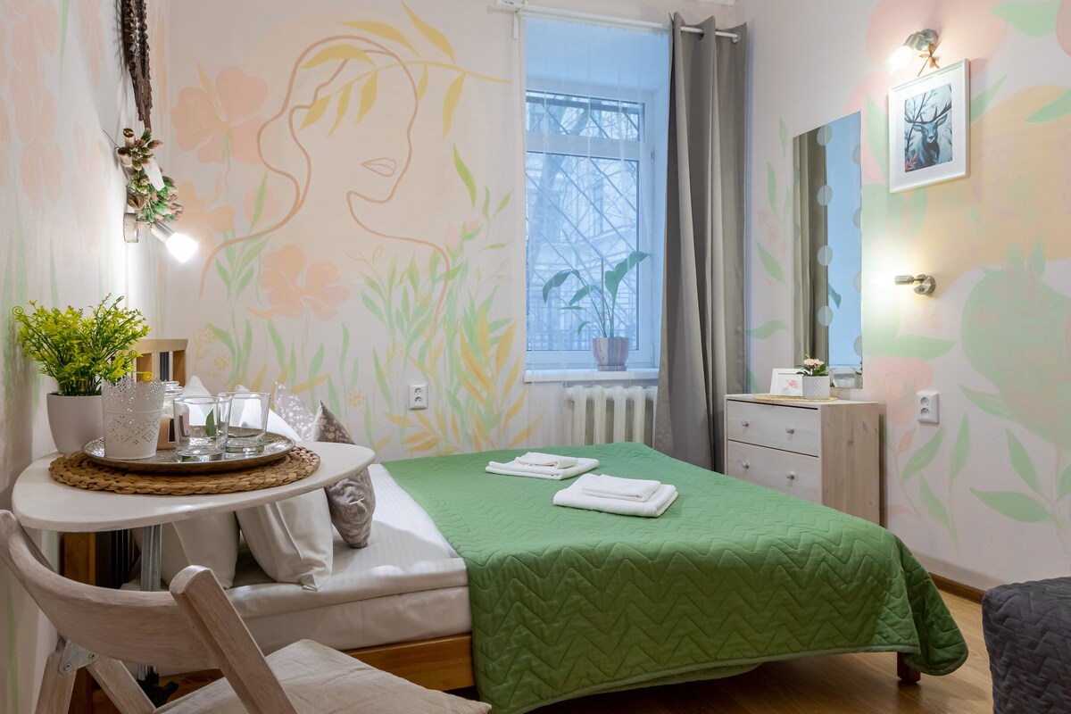 Une chambre avec une couvre-lit verte, une table de chevet ornée d’une plante, une fenêtre donnant sur un beau paysage et un mur décoré d’œuvres d’art florales.