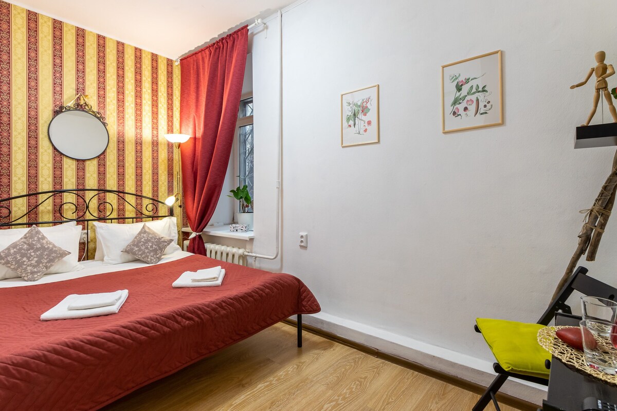 Un lit bien fait, recouvert d’une couverture rouge, une table de chevet avec une lampe, une chaise avec un coussin jaune et une fenêtre avec des rideaux rouges.