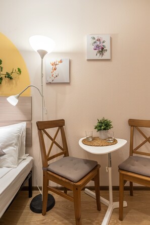 Une pièce bien rangée, avec un lit, deux chaises en bois, une petite table ornée d’une plante et deux lampes fixées au mur.