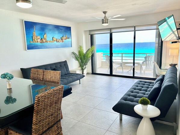 A modern living room with a large window overlooking the ocean, a ceiling fan, and a painting of a cityscape. - Sparkling Ocean Views at Villas Marlin (Cancun)