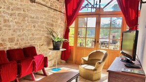 A cozy living room with a stone wall, red curtains, a wooden bench, a beige armchair, and a television on a wooden stand. - Gite du Coustal, Ancienne Grange, Cajarc, Lot (Cajarc)