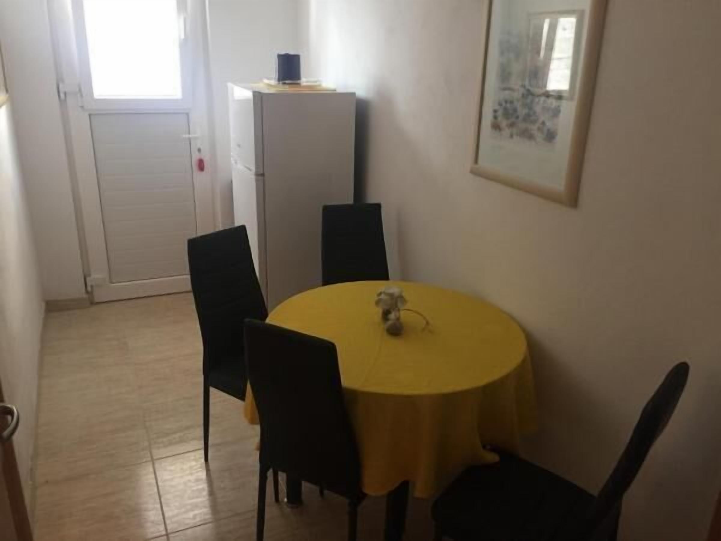 A small dining area with a round yellow table, four black chairs, a white refrigerator, and a framed picture on the wall.